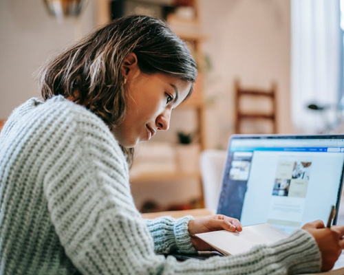a photo of a student with a laptop in front, writing on a notepad