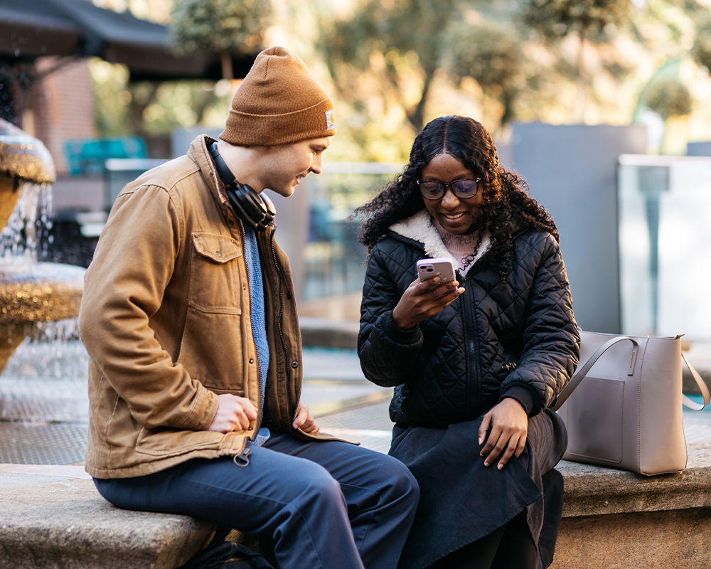 a photo of 2 students talking with one student looking at their phone